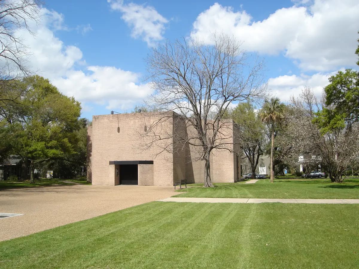 A windowless octagonal brick chapel with a dark entrance, set behind a wide green lawn and gravel plaza with bare and leafy trees under a blue sky