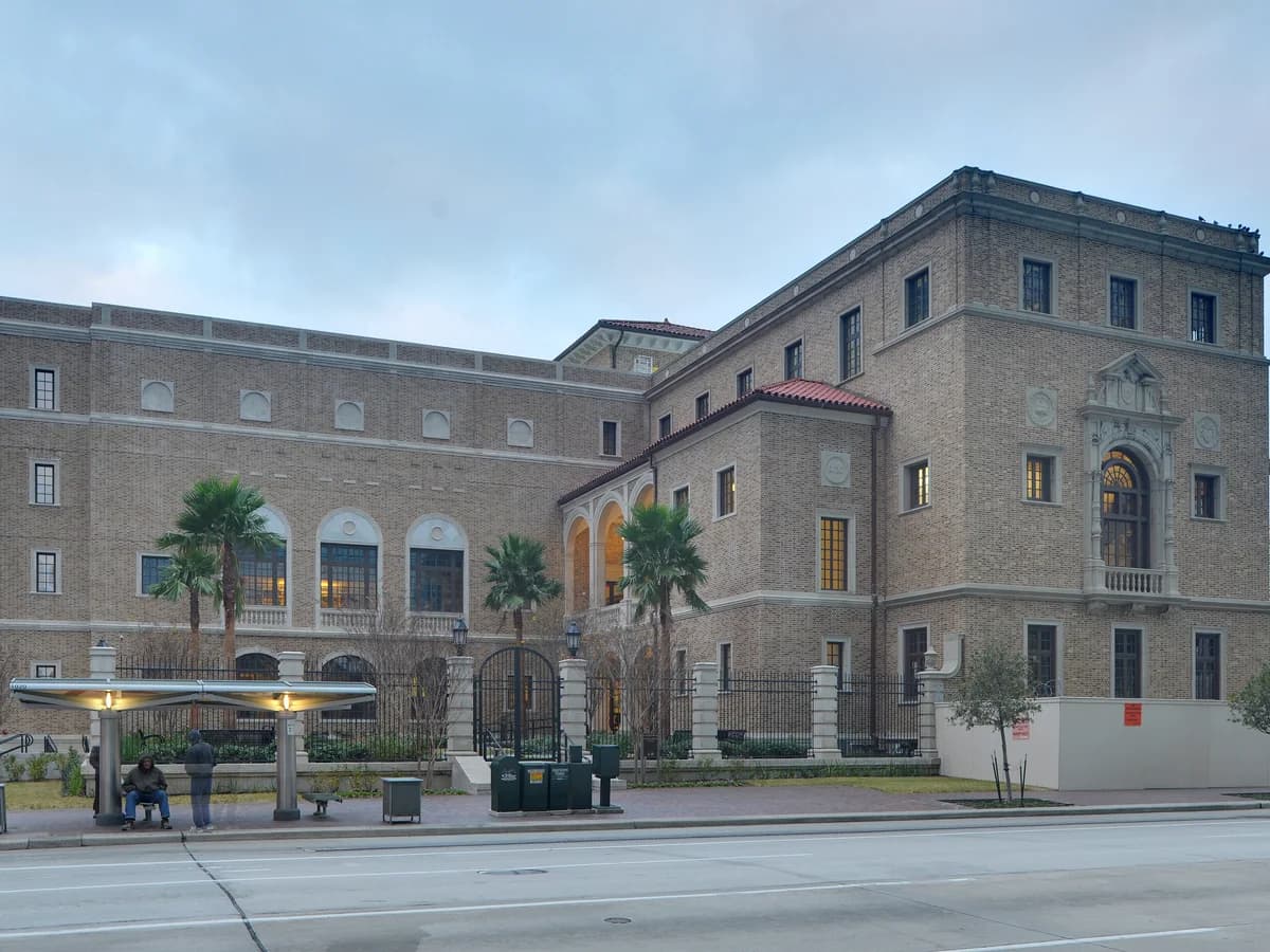 A tan brick Spanish Renaissance style library with arched windows, a red tile roof and palm trees, seen across a wide downtown street under a gray sky