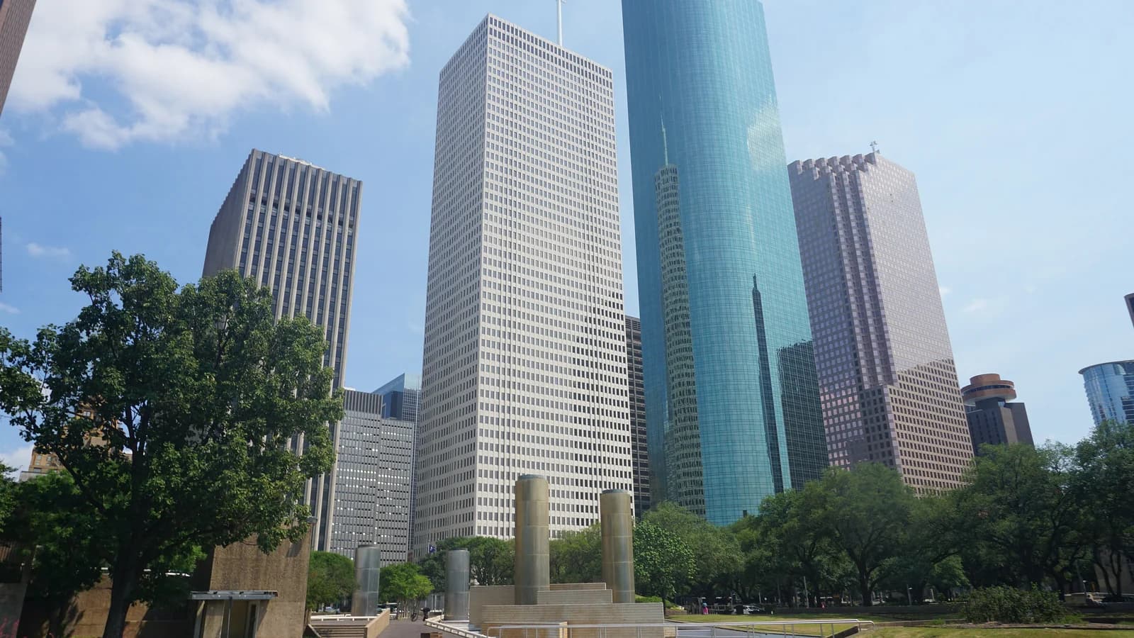 Downtown Houston towers, including a tall blue green glass skyscraper, rising behind the trees, lawns and short stone towers of Tranquillity Park on a sunny day