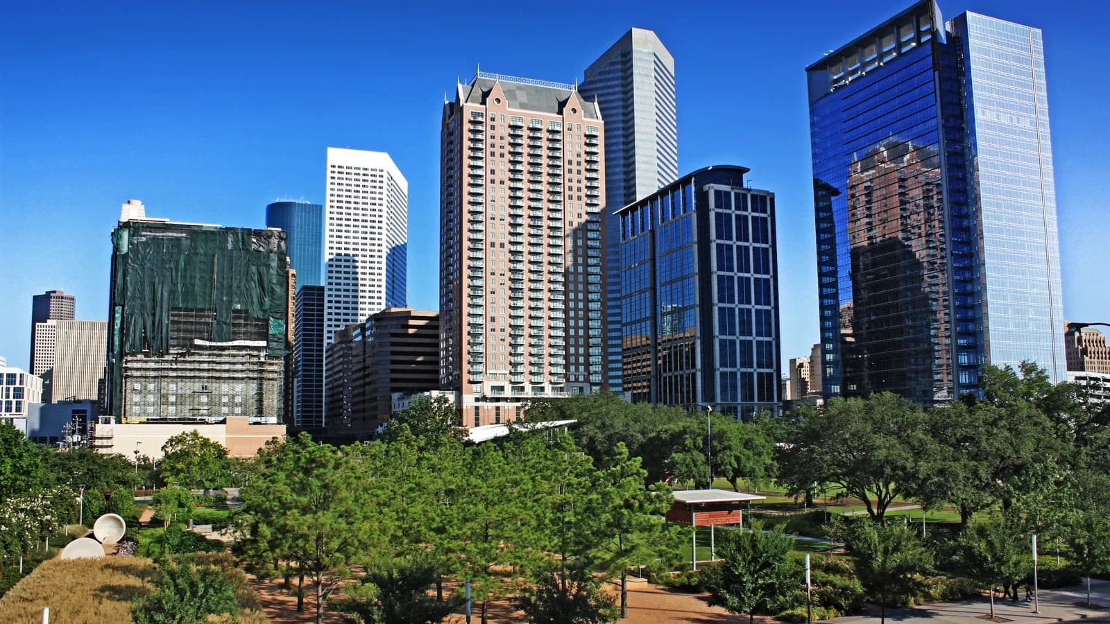 Discovery Green's leafy trees and paths below a cluster of downtown Houston glass and stone towers under a deep blue sky