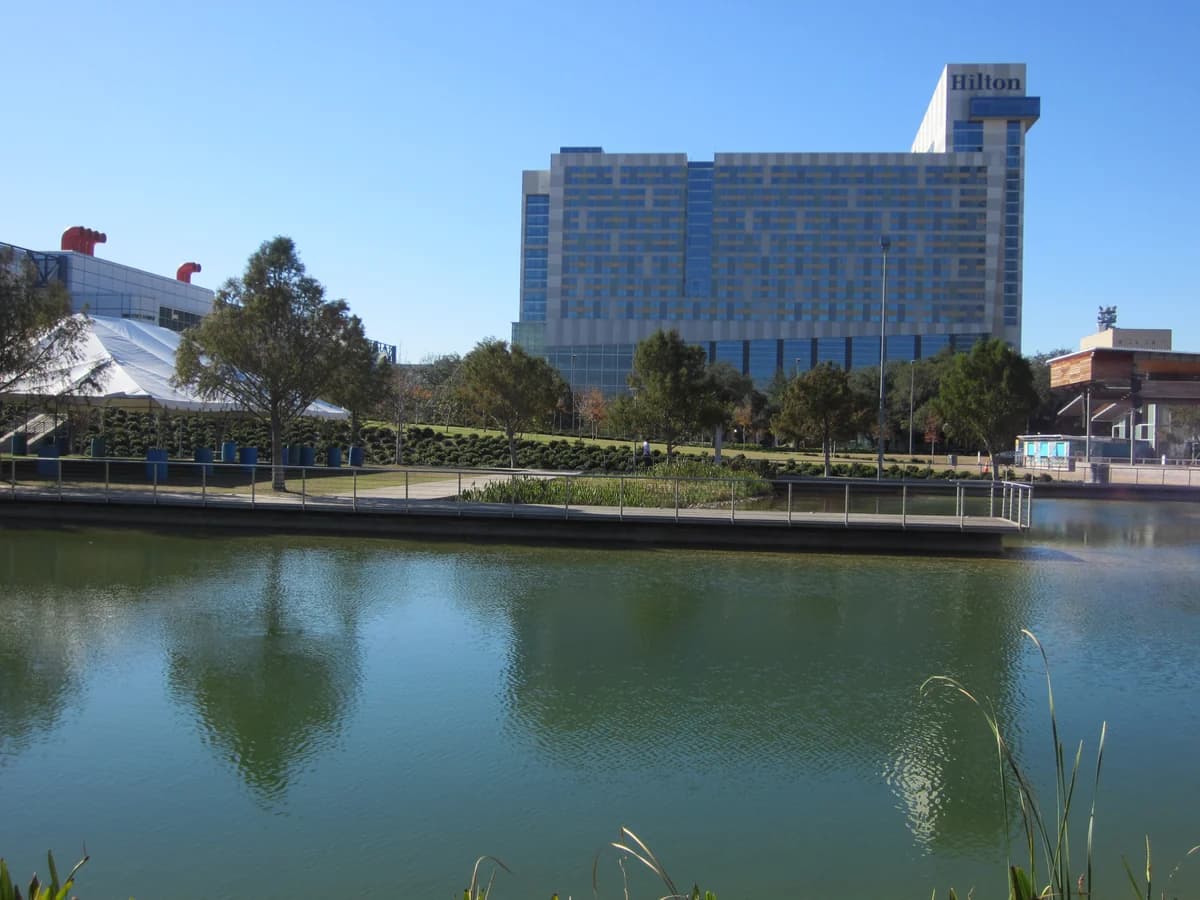 Kinder Lake at Discovery Green reflects the trees on its far bank, with the tall Hilton Americas hotel tower and a white tent roof behind
