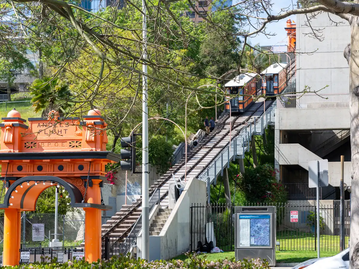 An ornate orange arch reading Angels Flight at the foot of a steep funicular track, with two orange and black cars passing high up among trees and concrete buildings