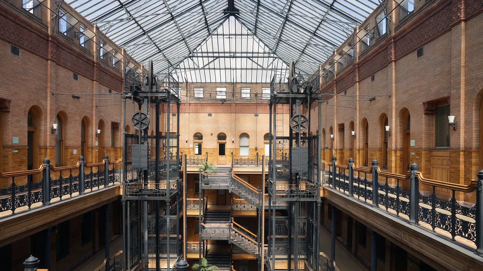 The sunlit atrium of the Bradbury Building, with ornate black iron railings, open cage elevators and a glass roof