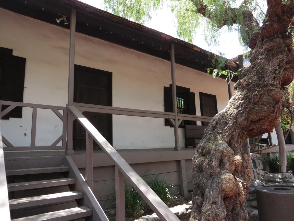 Whitewashed adobe house with a shaded wooden veranda, railed steps and dark shuttered windows beside a gnarled old pepper tree