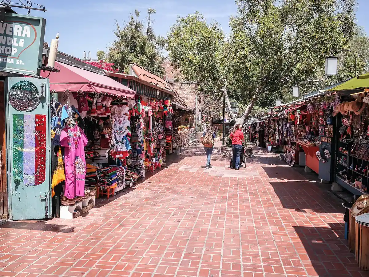 A wide red brick lane lined with market stalls hung with embroidered dresses, bags and souvenirs under awnings and big shade trees, with a few people walking away