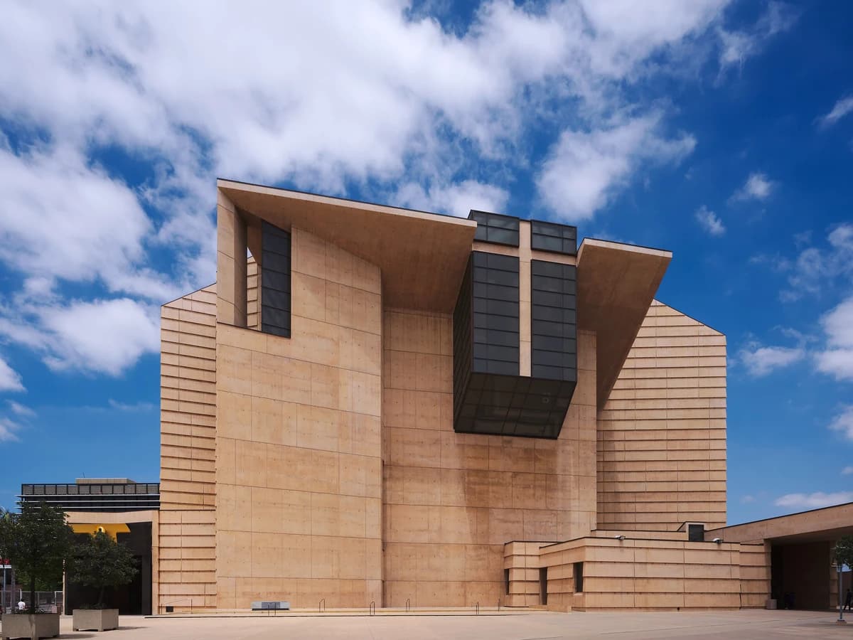Angular ochre concrete cathedral facade with a cross-shaped window of dark glass, rising above an empty stone plaza under a blue sky with clouds