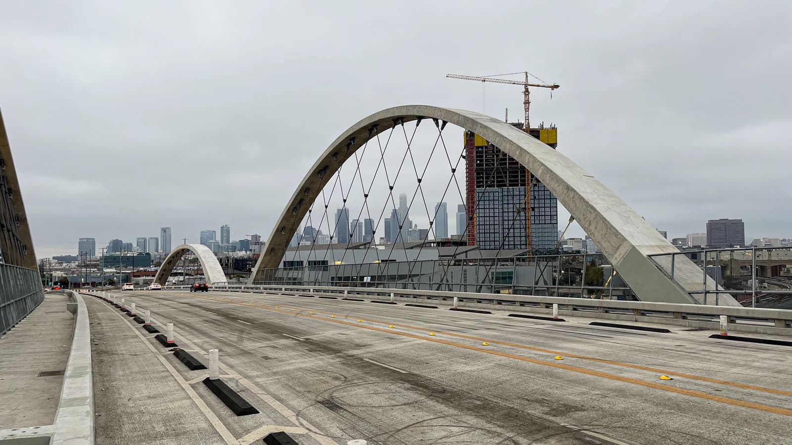 A pale concrete arch of the Sixth Street Viaduct rising over its empty deck, with the downtown Los Angeles skyline under an overcast sky behind
