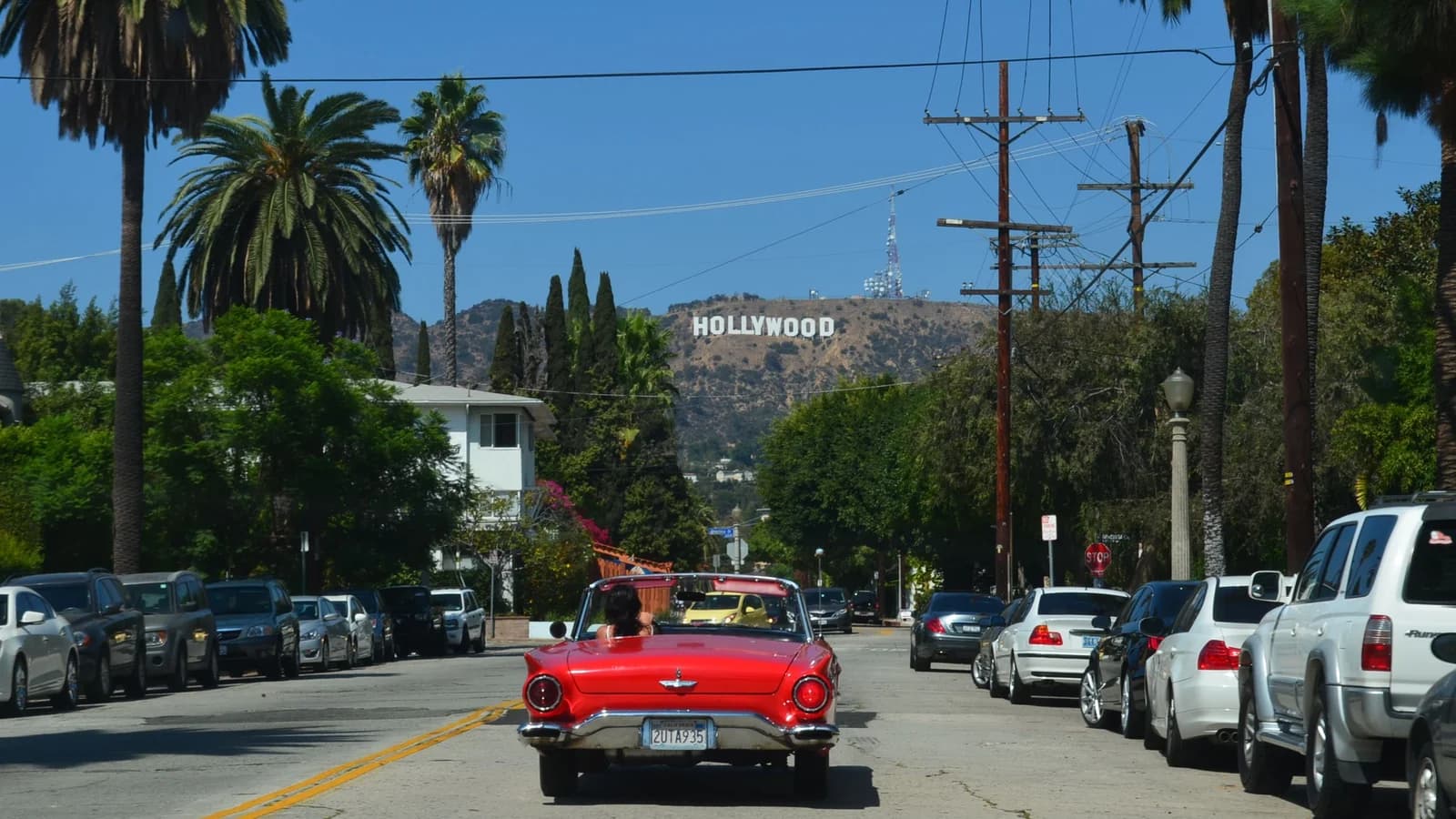 A red vintage convertible drives up a palm-lined Hollywood street with parked cars, and the Hollywood Sign on the hills ahead under a blue sky