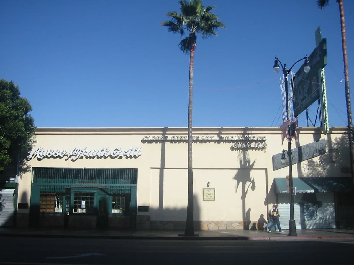 Cream storefront with script Musso & Frank Grill lettering, green trim and a tall roadside sign under palm trees on a sunny boulevard