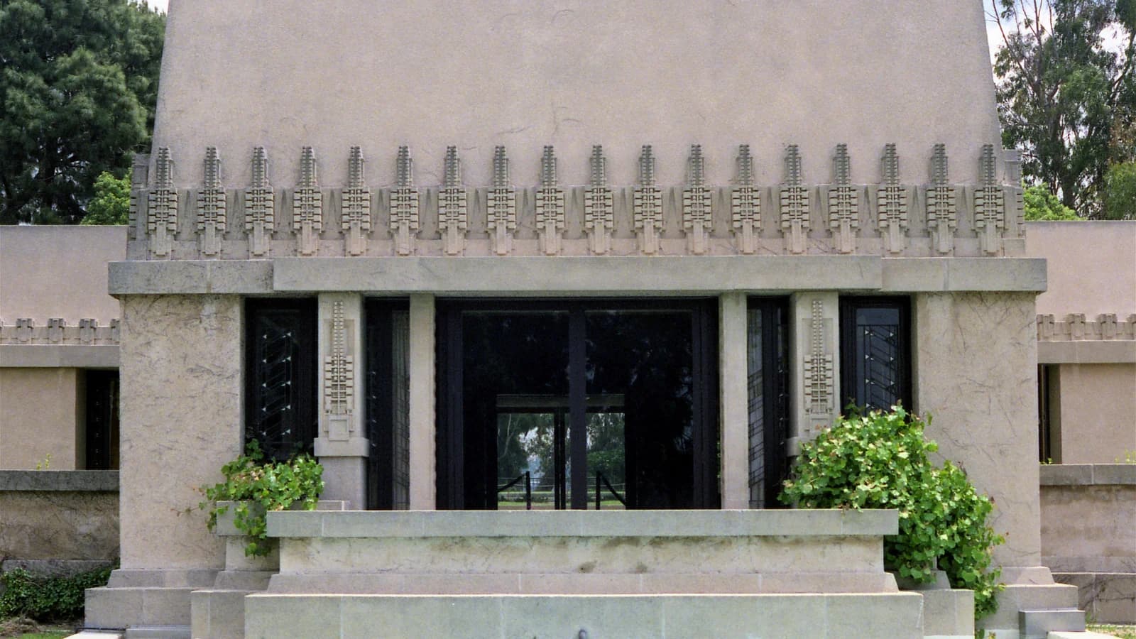 The cream colored concrete Hollyhock House by Frank Lloyd Wright, with a row of stylized hollyhock ornaments above tall dark windows and a low concrete basin in front