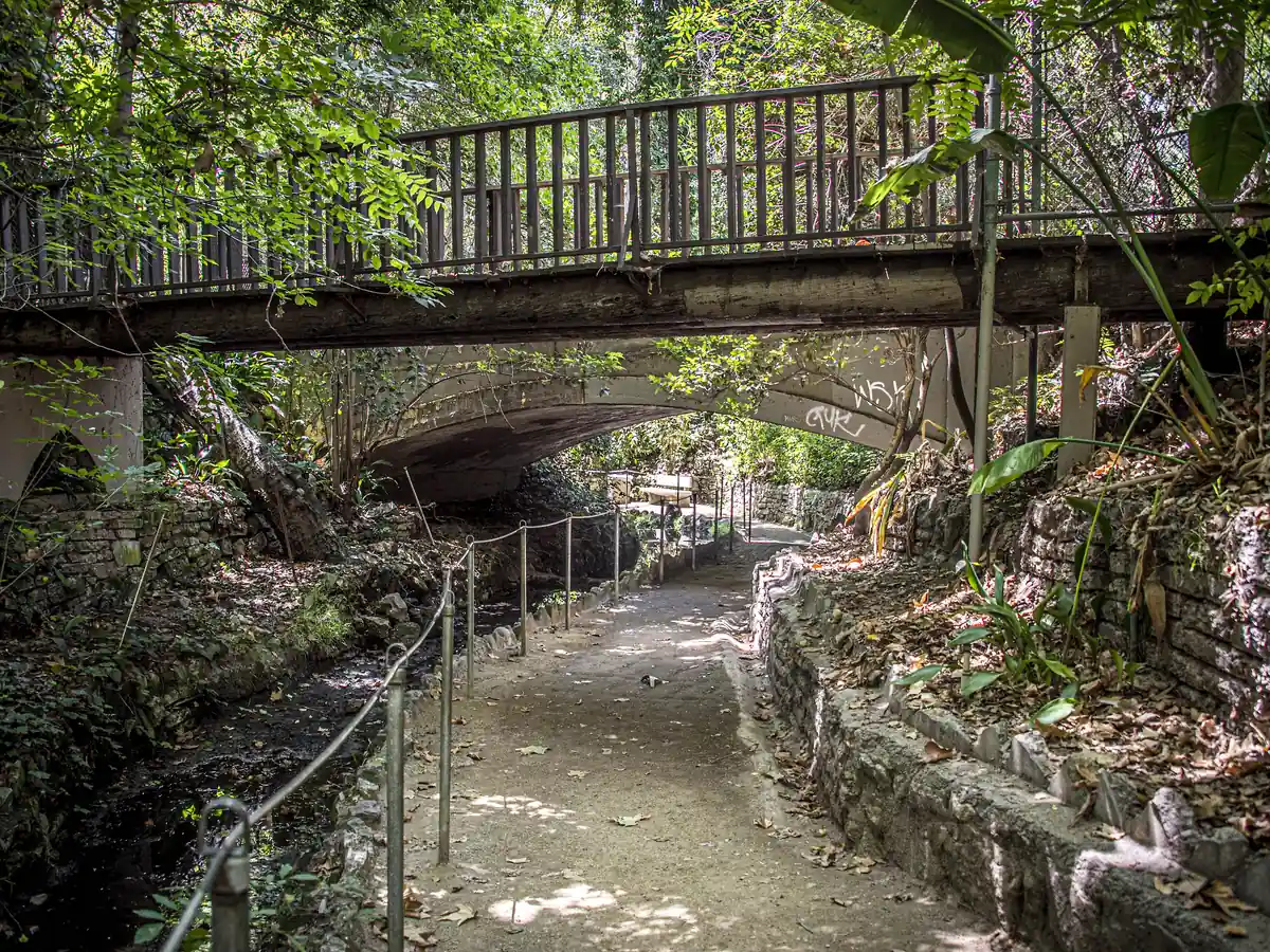 Shaded dirt path with a metal handrail beside a small stream, running under a railed footbridge and an old arched bridge among ferns and trees