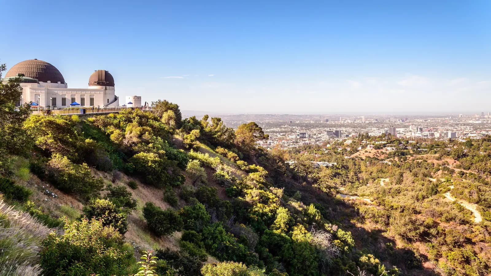 The white art deco Griffith Observatory and its copper domes atop a green, scrubby hillside, with the Los Angeles basin stretching to the horizon