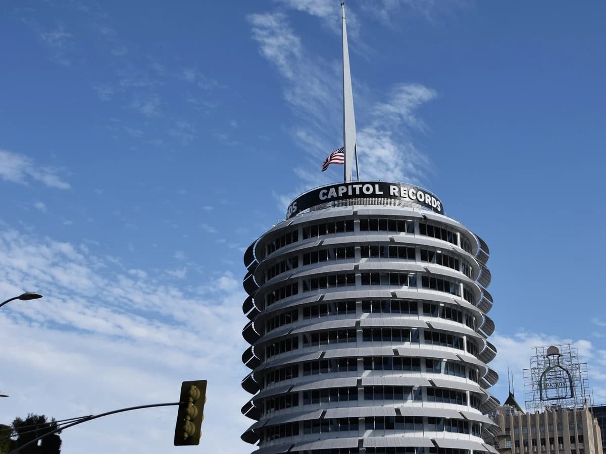 Round white tower with stacked curved floors, a Capitol Records sign, a flag and a tall spire on top, under a blue sky with thin clouds