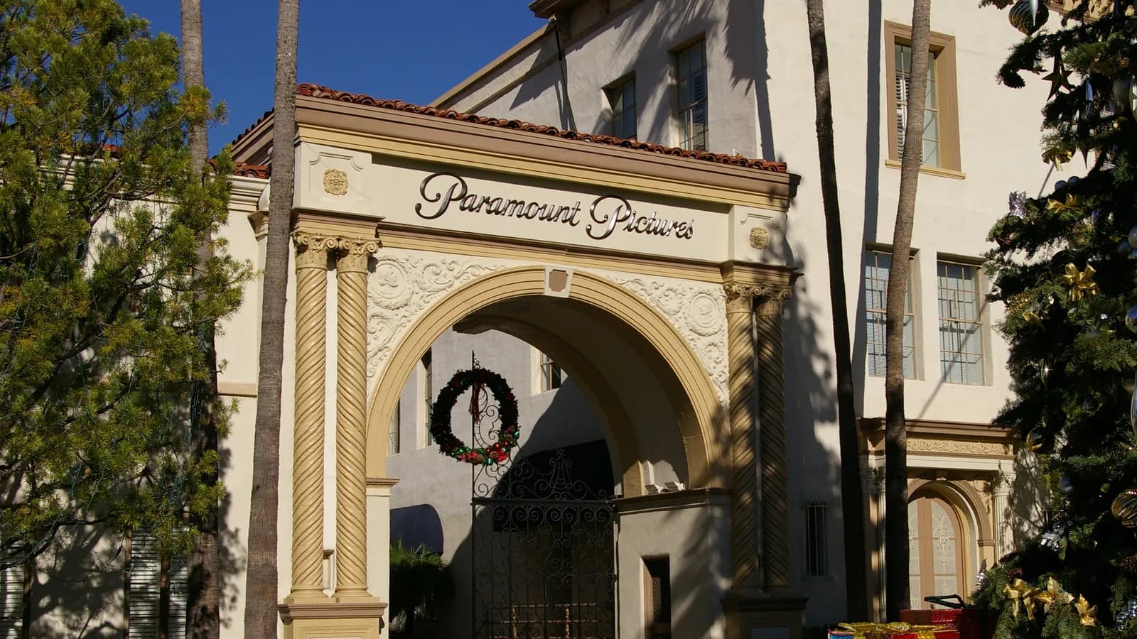 The ornate cream arch of Paramount's Bronson Gate with Paramount Pictures lettering, a holiday wreath on its iron gate, palms and a decorated tree beside it