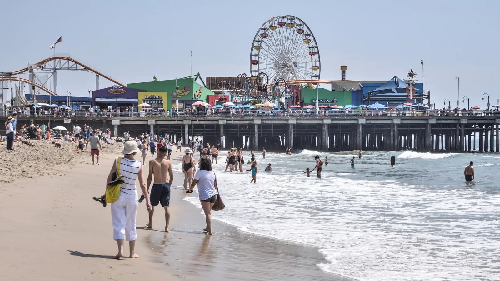 The Santa Monica Pier with its Ferris wheel and roller coaster seen from the beach, with surf rolling in and people strolling along the sand