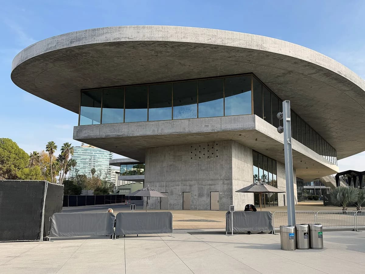 A wide curved concrete roof over a band of tall windows, raised on a bare concrete pier above a plaza with umbrellas, with palm trees and a glass tower behind