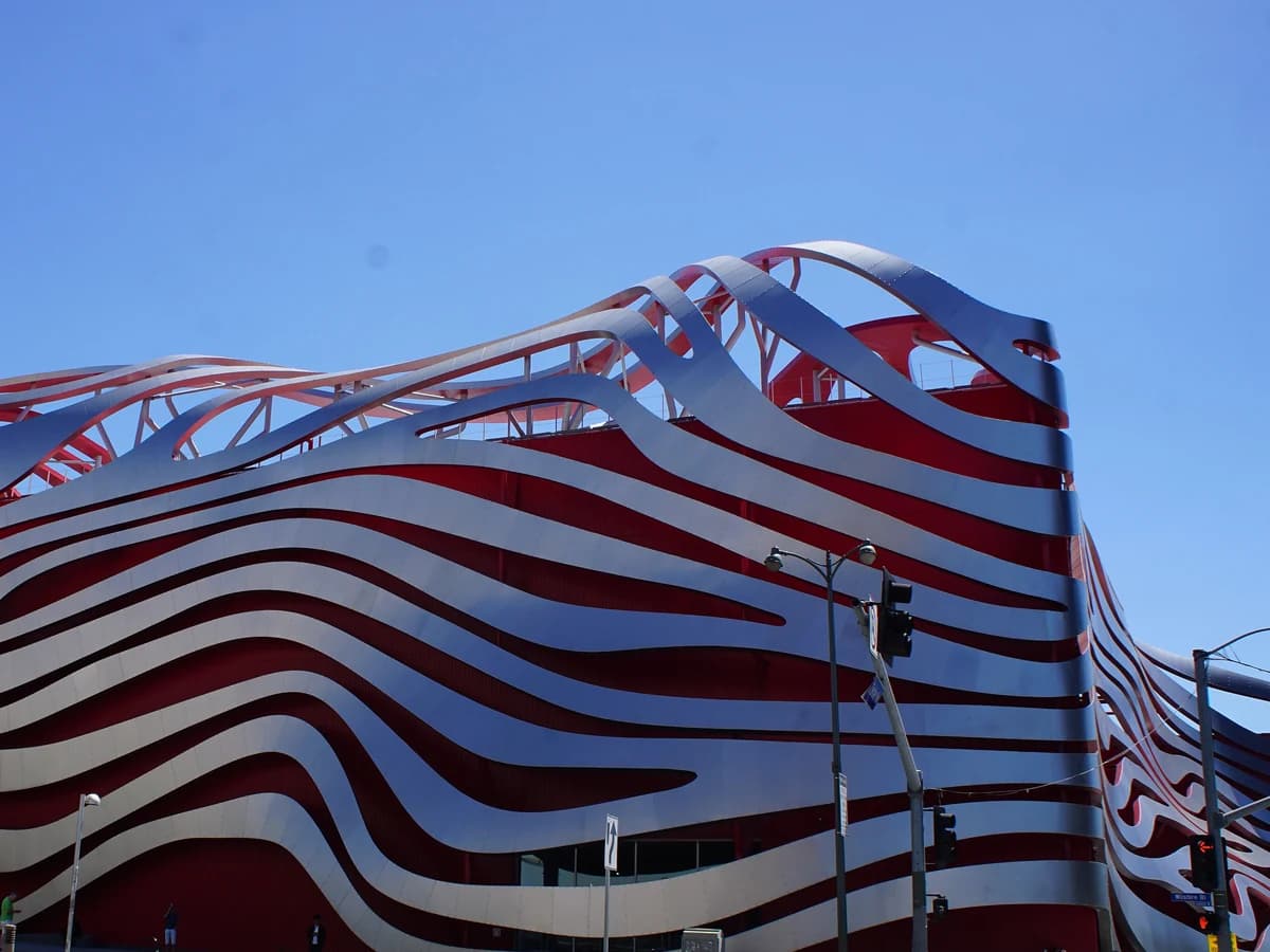 A building wrapped in wavy ribbons of brushed stainless steel over a bright red facade, with a street light and traffic signal in front under a clear blue sky