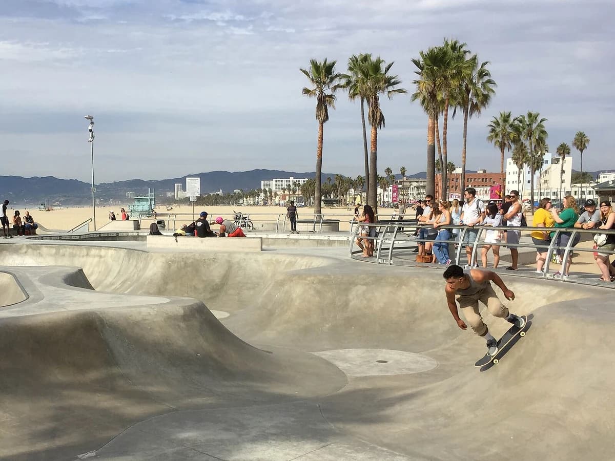 A skateboarder carving through a smooth concrete bowl, with onlookers at the rail, tall palm trees, the beach and hazy mountains behind