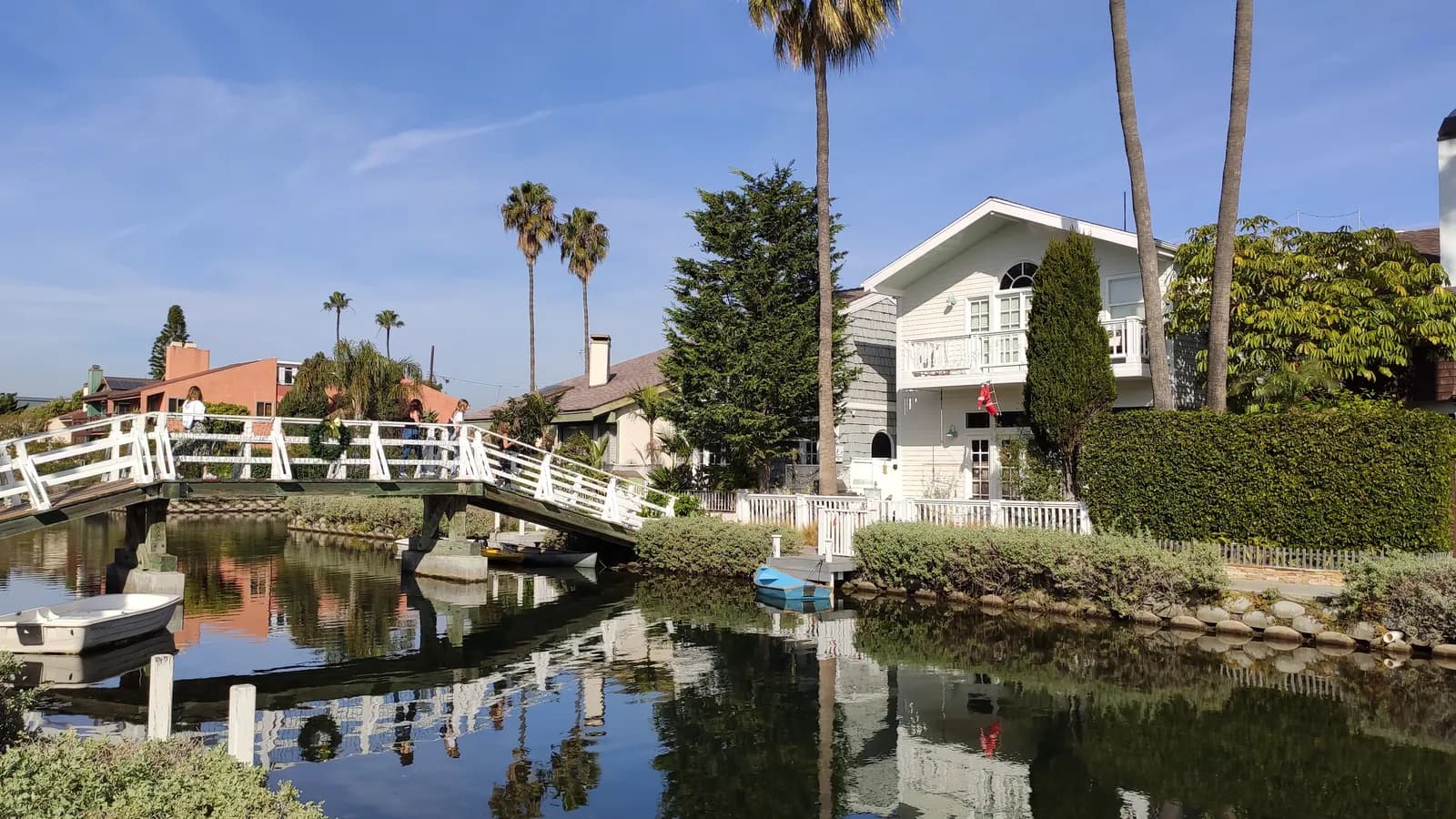 A white footbridge arching over a calm Venice canal, with small boats, hedges, tall palms and pastel houses reflected in the water under a blue sky