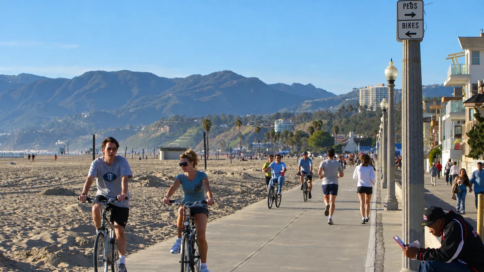 A runner in a gray shirt on the Santa Monica beach path among cyclists and walkers, with sand on the left and the Santa Monica Mountains behind