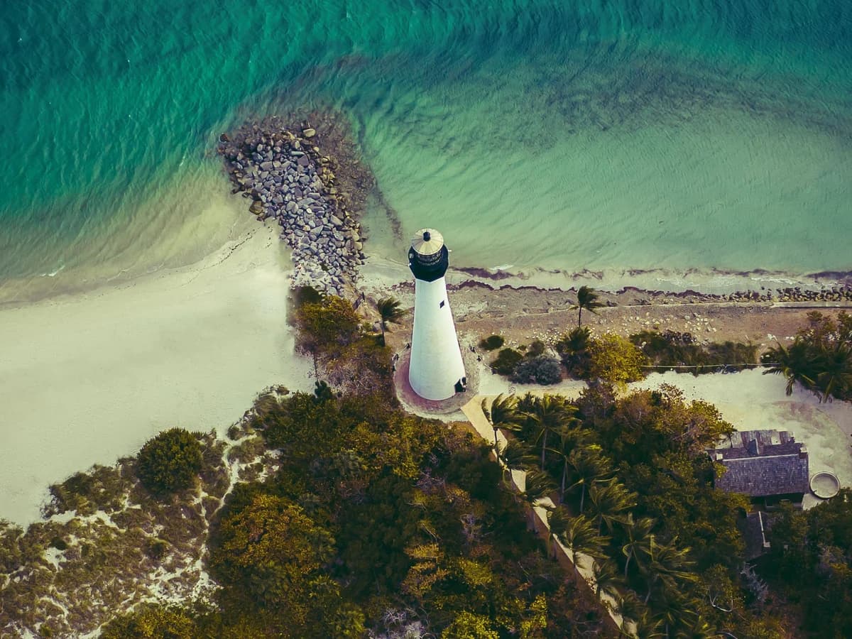Aerial view of a white lighthouse with a black lantern beside a rock jetty, where a pale sand beach meets turquoise water, with dense green trees and palms below