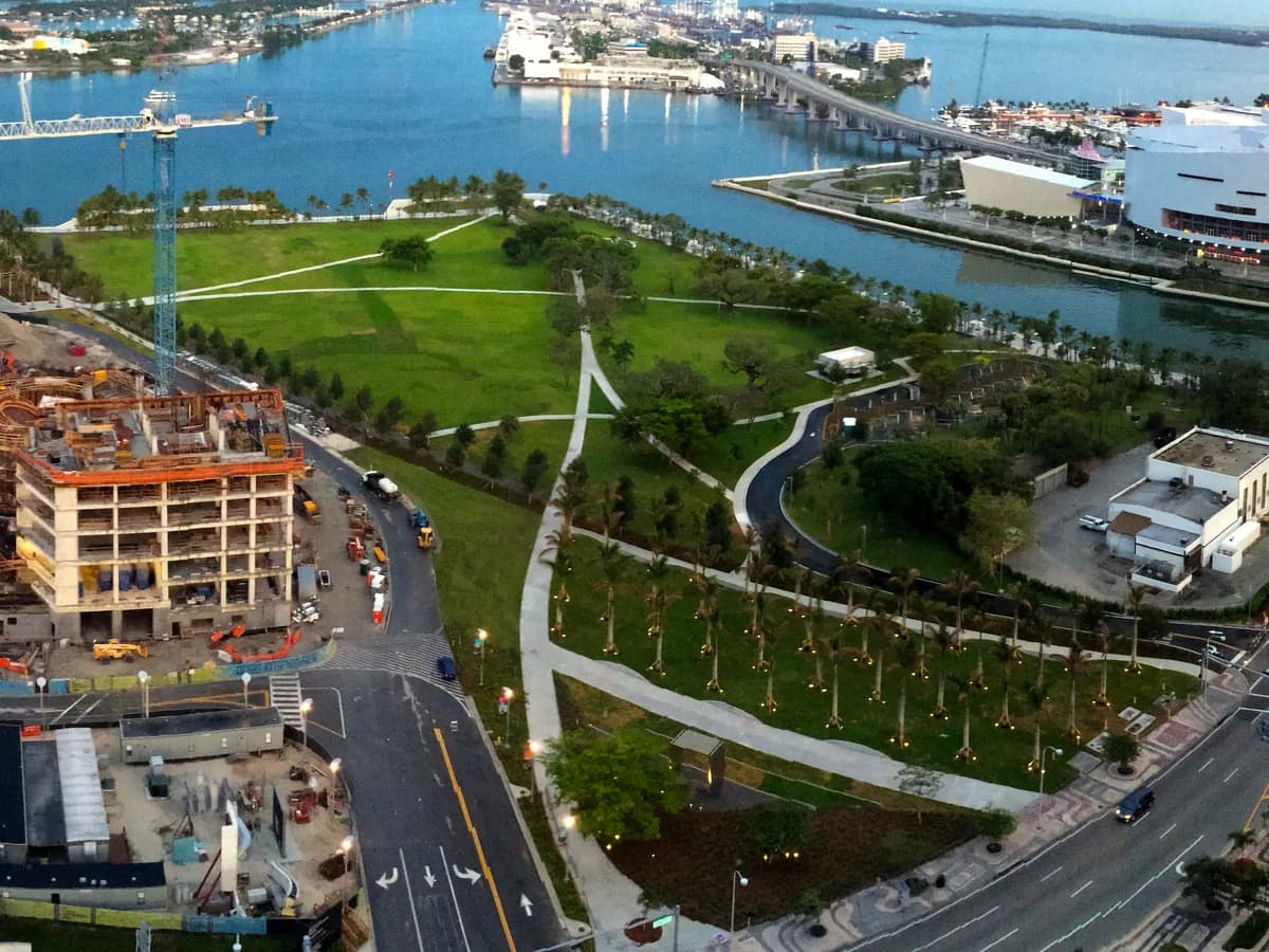 A high view over a bayfront park with wide lawns, rows of young palms and curving paths, with Biscayne Bay, a round white arena and towers beyond and a building site at the left