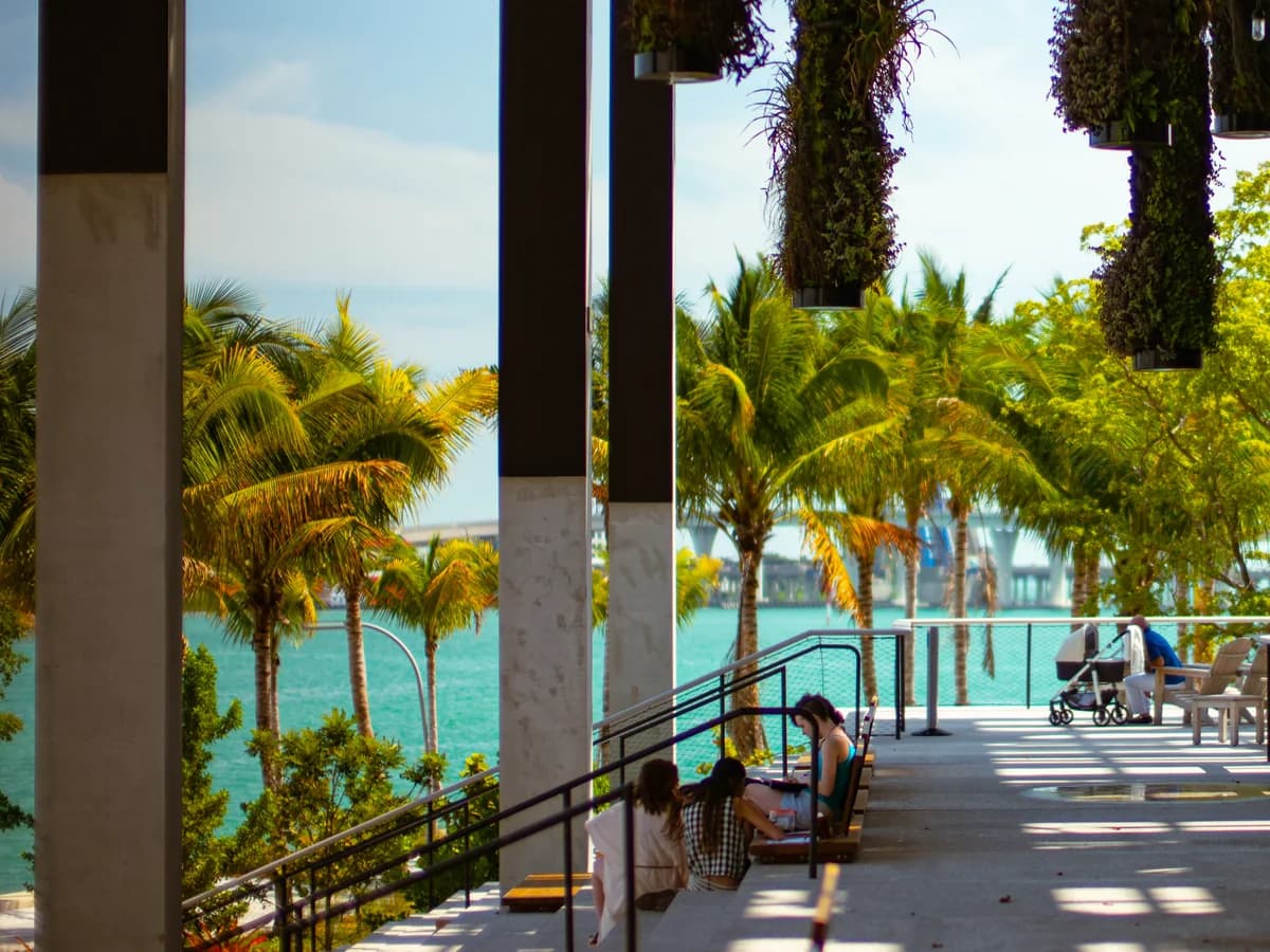 A shaded museum veranda with slender concrete columns and hanging plants, looking out past palm trees to the turquoise water of Biscayne Bay, with visitors seated by the railing