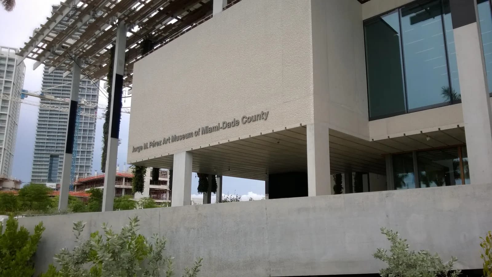 A modern concrete museum raised on slender columns under a wooden canopy, with hanging plants, palm trees and glass towers behind