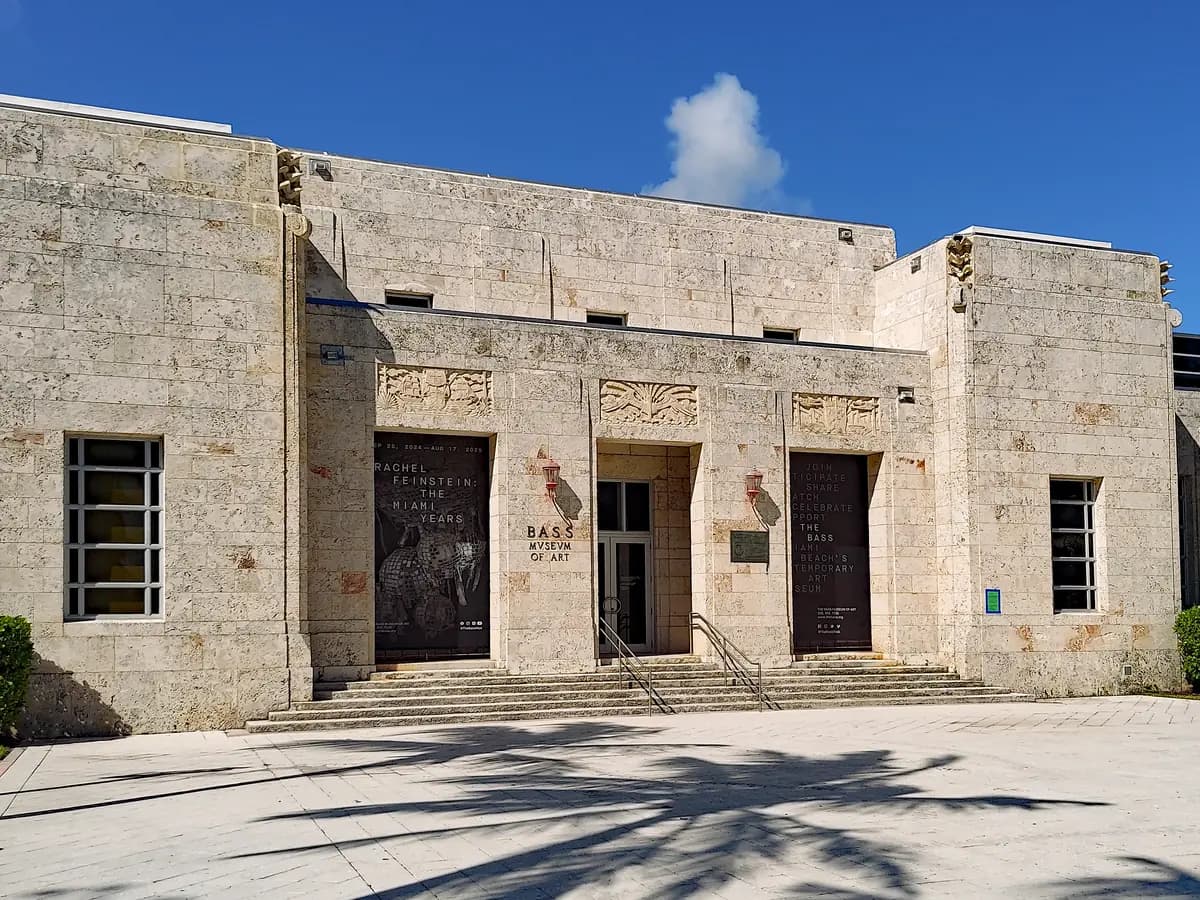 The pale coral stone front of the Bass museum with dark double doors, carved panels over the entrance and wide steps, and palm shadows on the plaza