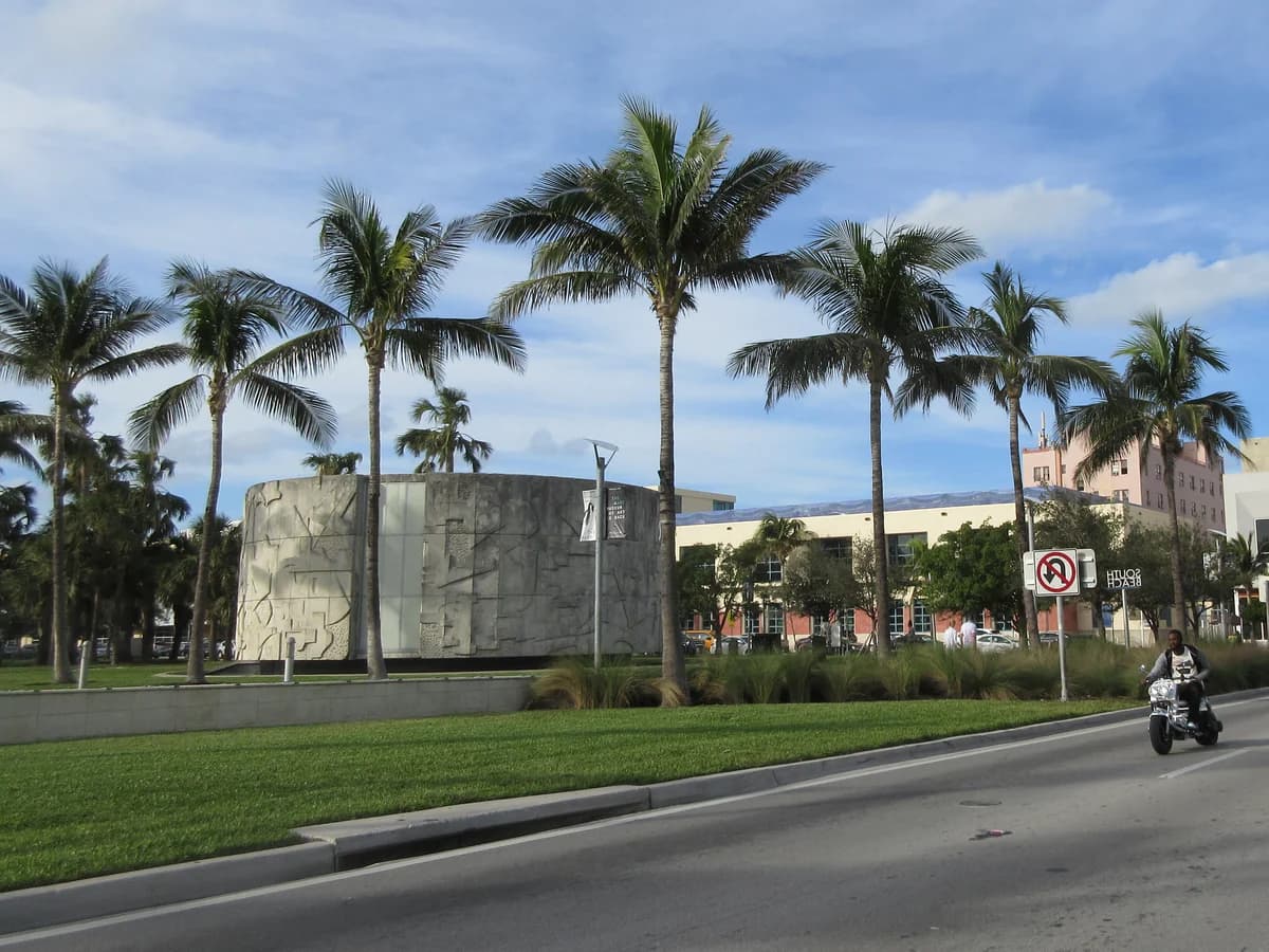 Coconut palms along a grassy park beside Collins Avenue, with a round stone rotunda, low buildings and a motorcyclist on the road under a partly cloudy sky