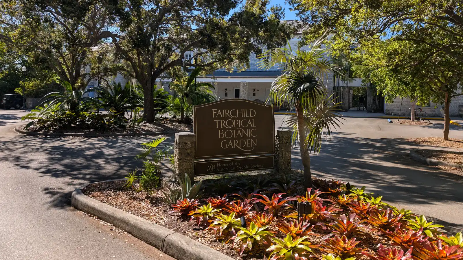A carved wooden sign for Fairchild Tropical Botanic Garden in a bed of red and green bromeliads, under large shade trees on a sunny day
