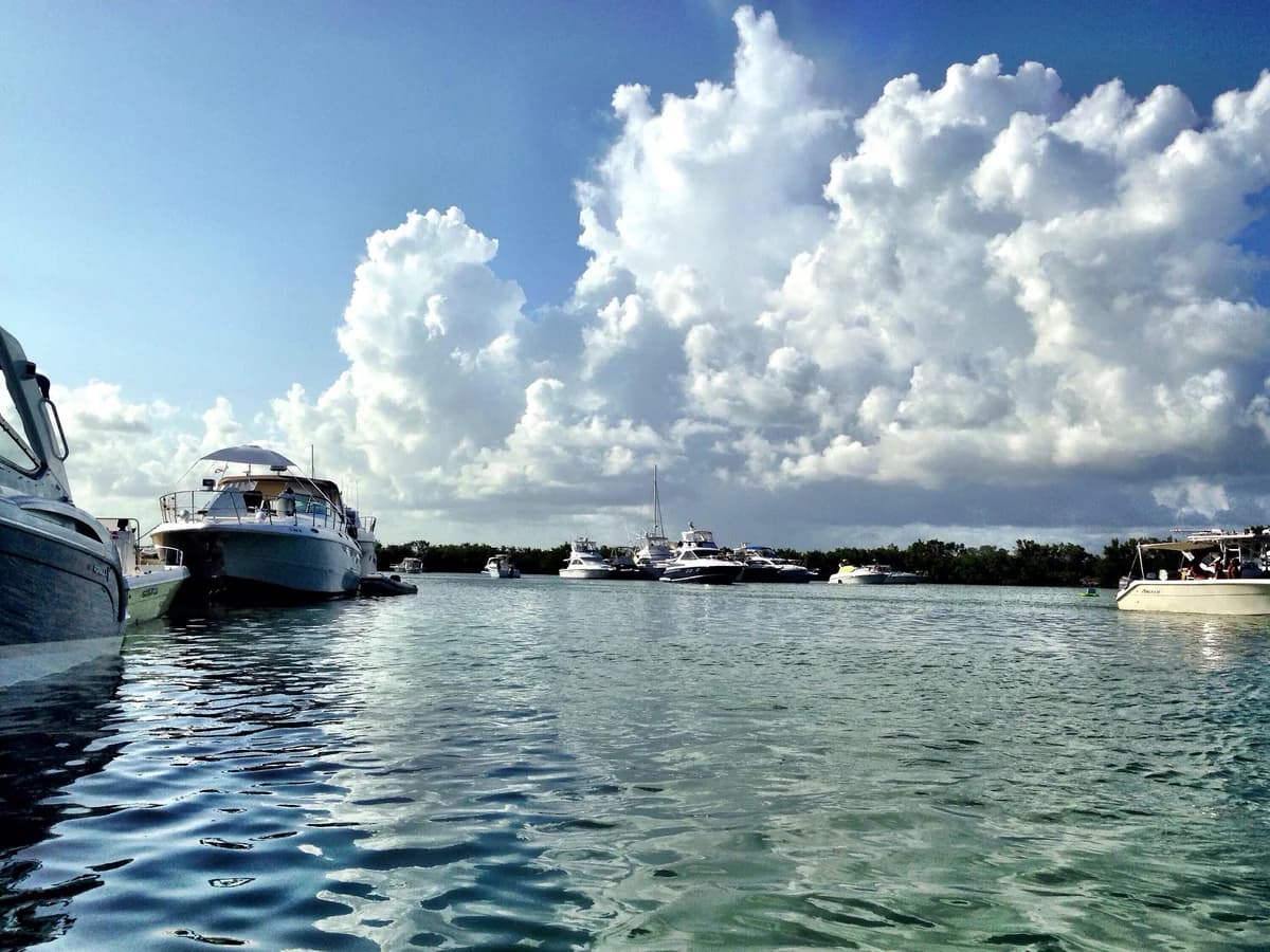 Motor yachts moored on calm green water in a sheltered harbor ringed by trees, under towering white clouds and a blue sky