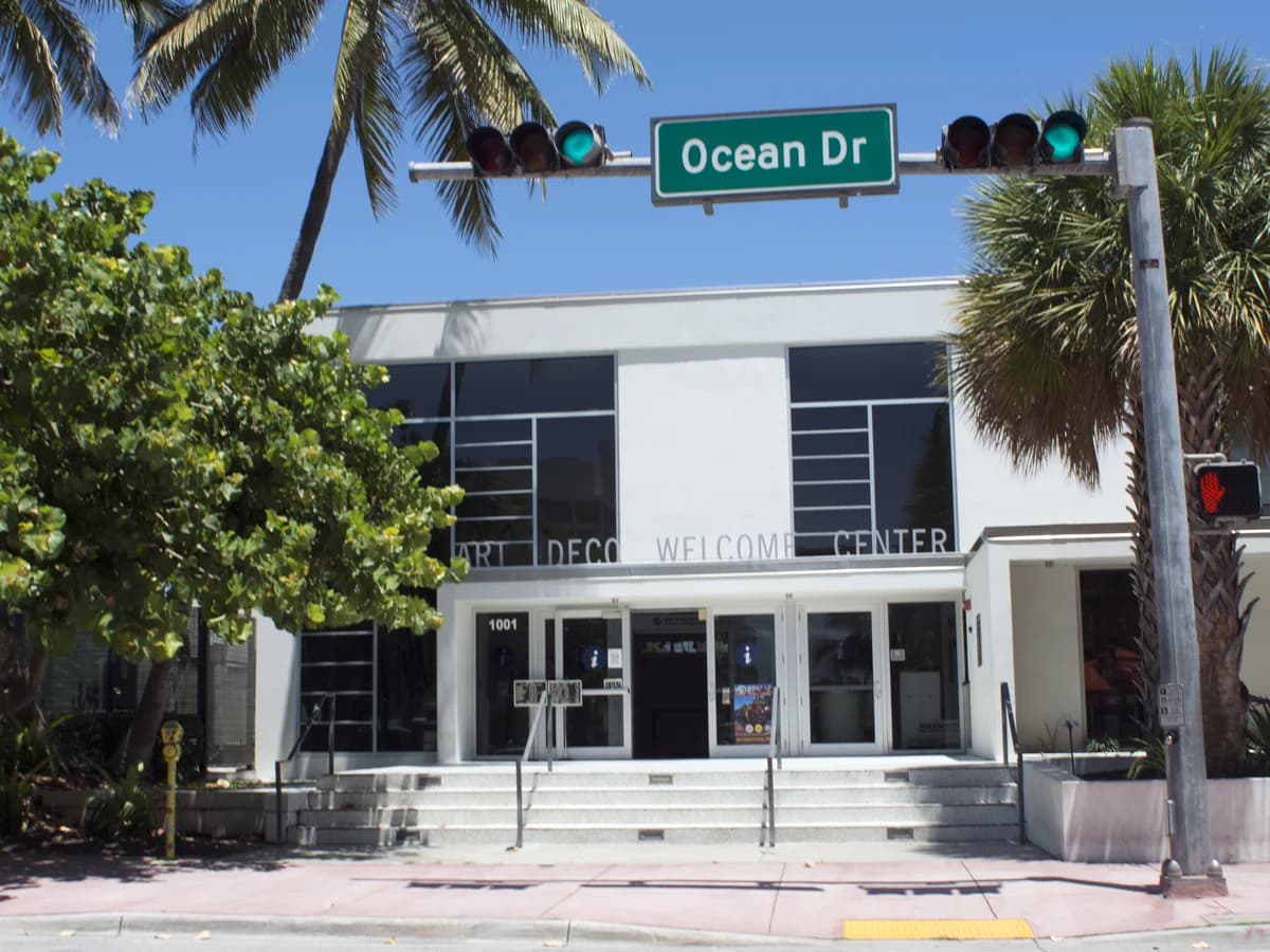 A white Art Deco building with tall dark windows and Art Deco Welcome Center lettering over the doors, under an Ocean Dr street sign and traffic lights, framed by palms and trees