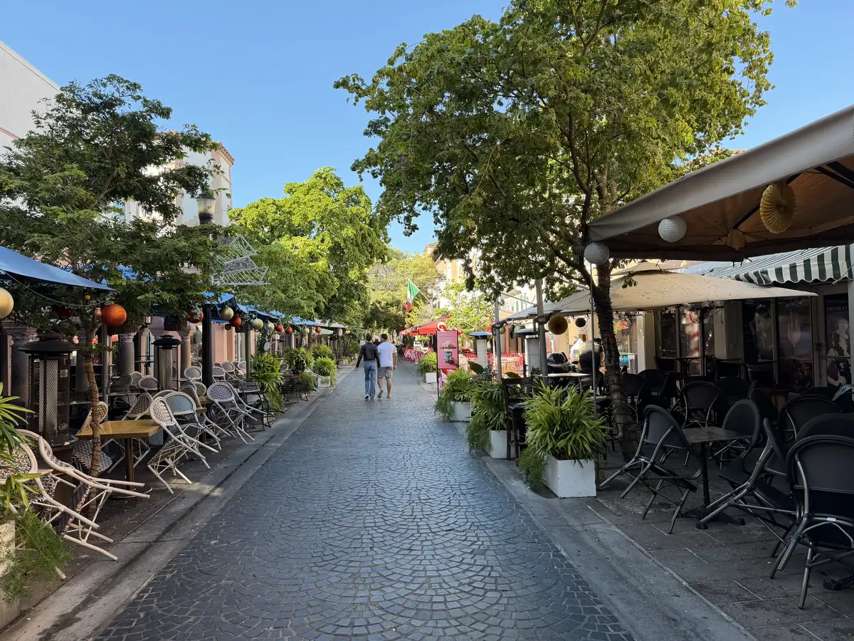 A narrow paved pedestrian street lined with cafe tables, umbrellas and potted plants, with leafy trees, pale buildings and a few walkers far down the middle