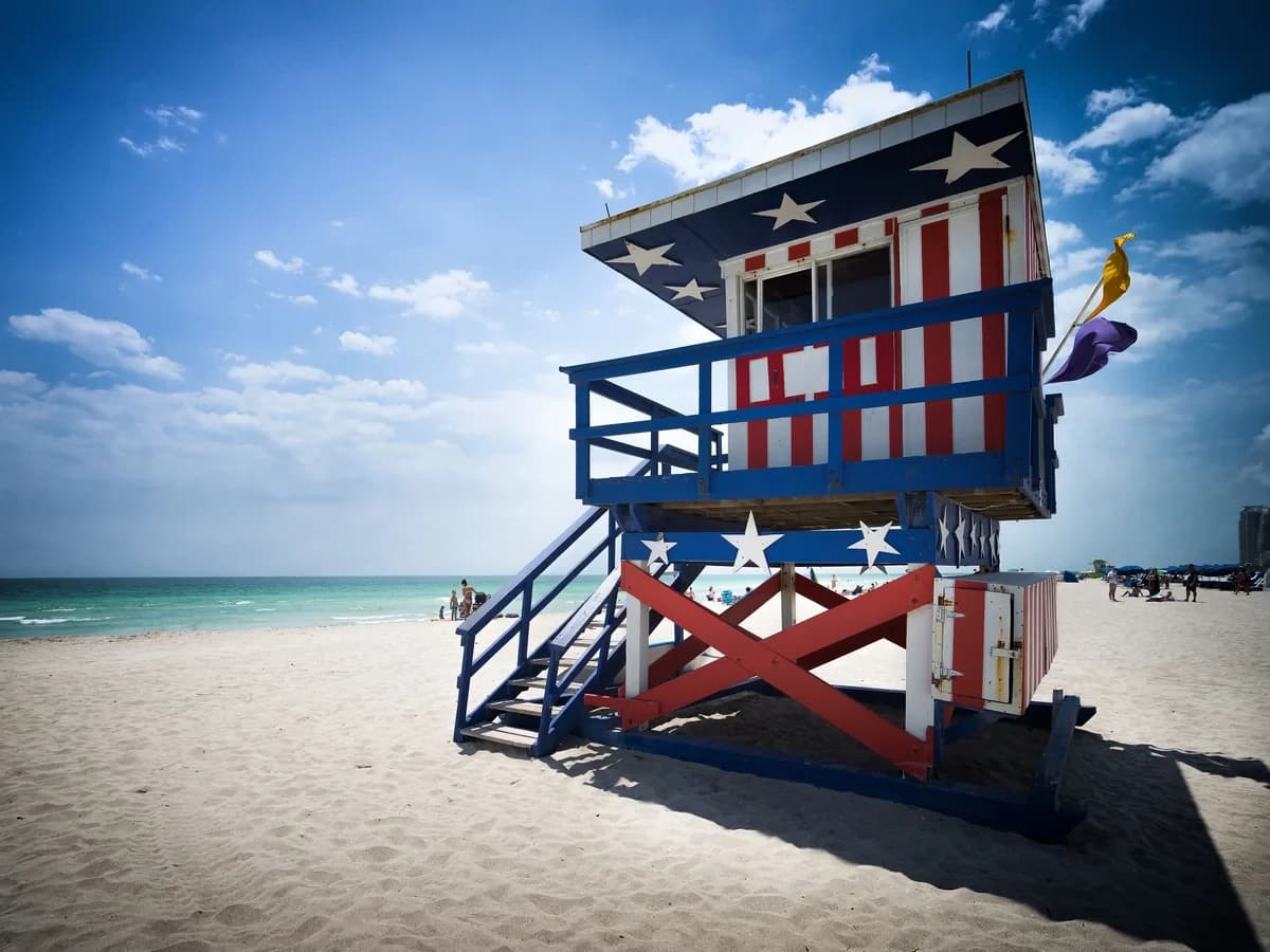 A lifeguard tower painted with stars and red and white stripes on white sand, with the turquoise ocean and a deep blue sky