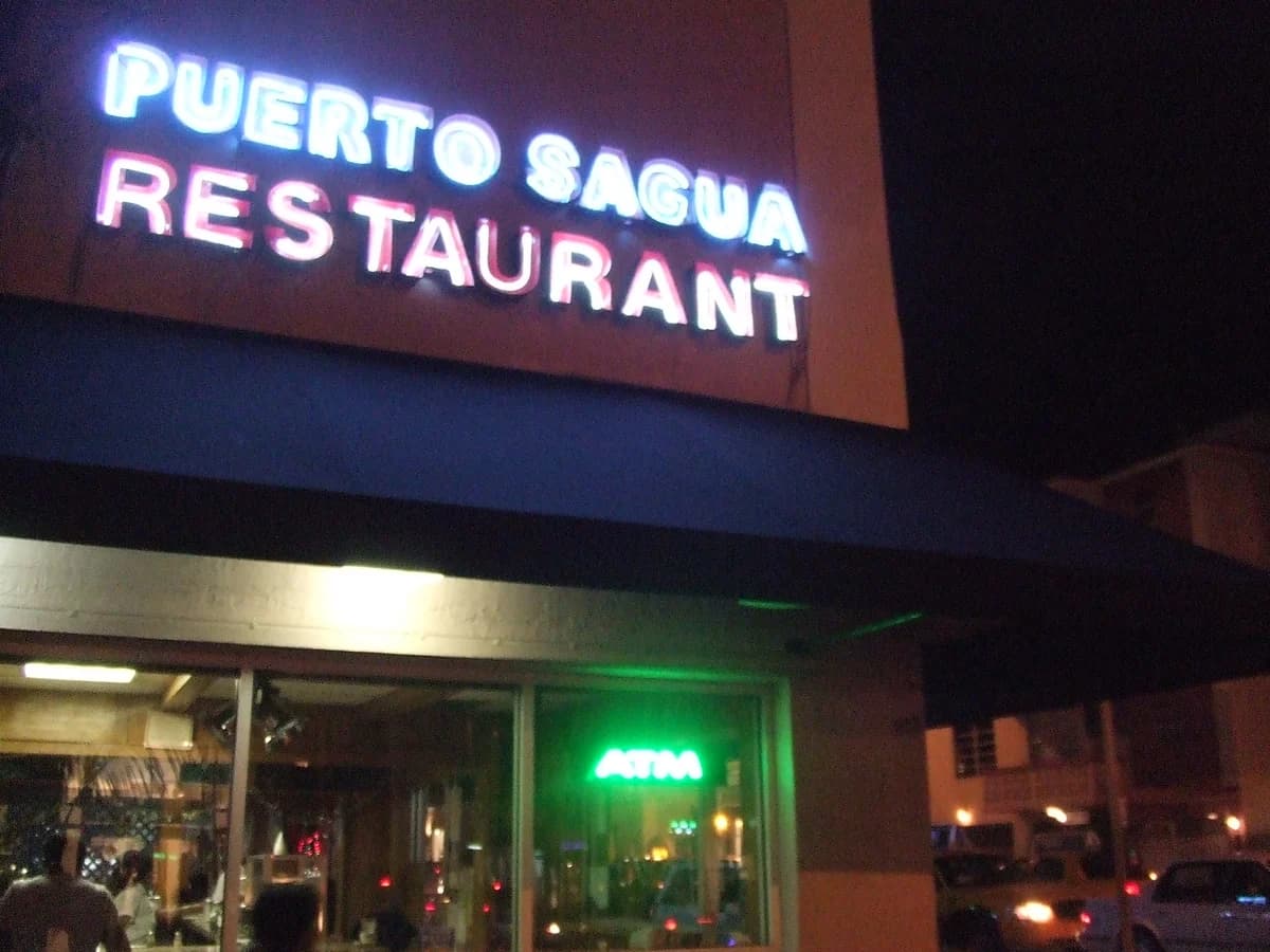 A glowing white neon sign reading Puerto Sagua Restaurant above the lit front windows at night, with cars on the street at the right