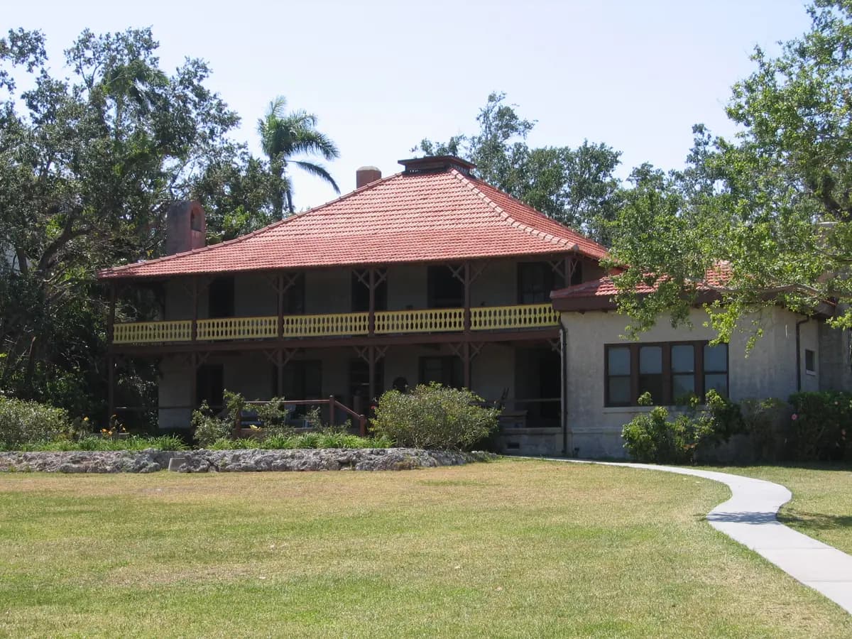 A wooden two story house with a wide red roof and a long upper porch on a green lawn, with a curving path, a palm and shade trees behind