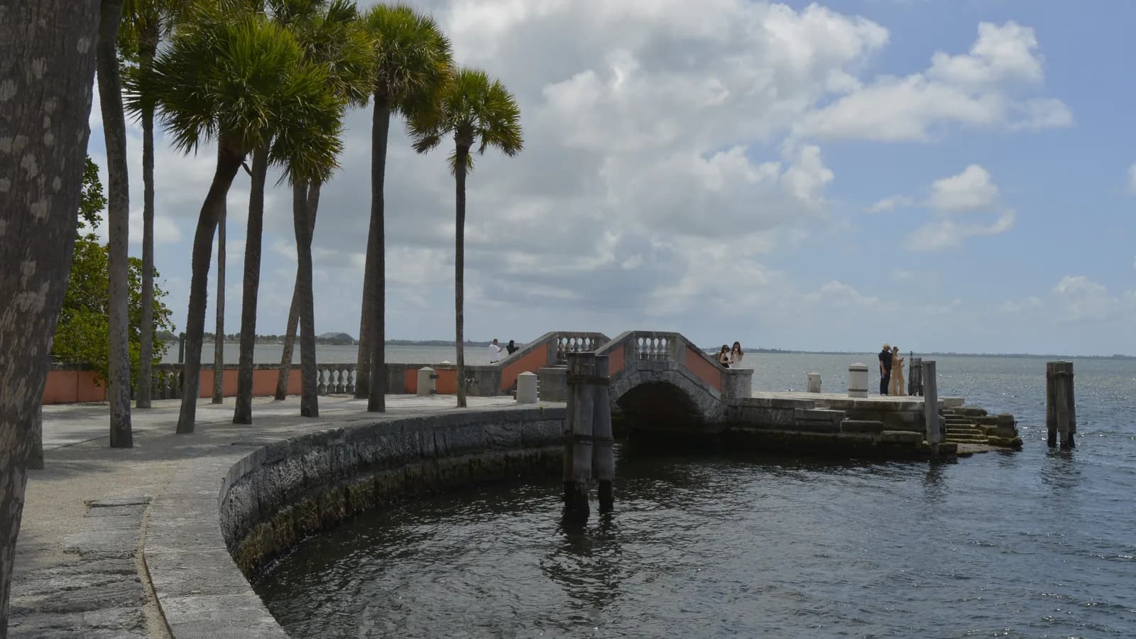 A curved stone seawall and a small arched stone bridge at the edge of Biscayne Bay, with tall palm trees and white clouds over the water