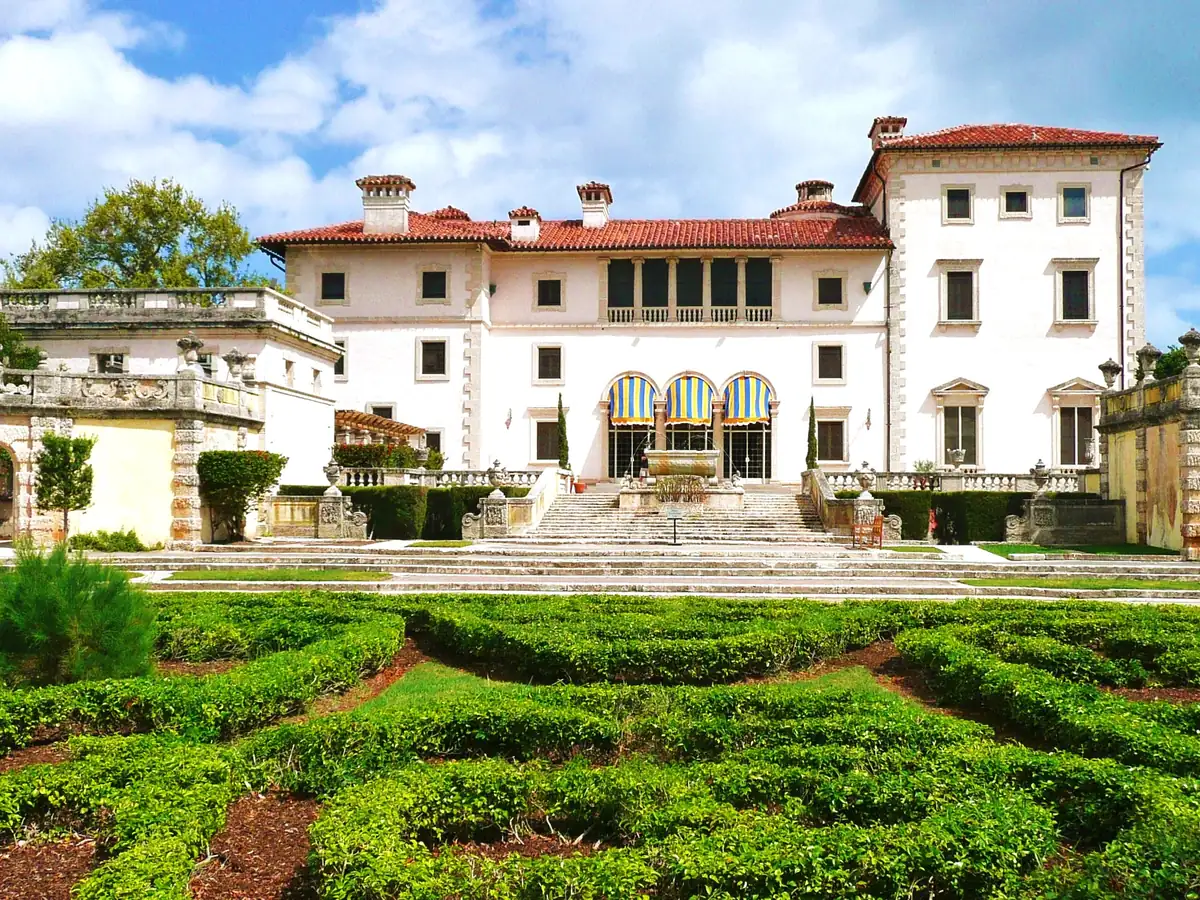 The cream stone Vizcaya main house with a red tile roof and arched windows, seen across clipped hedge parterres and a wide stone stairway under a blue sky