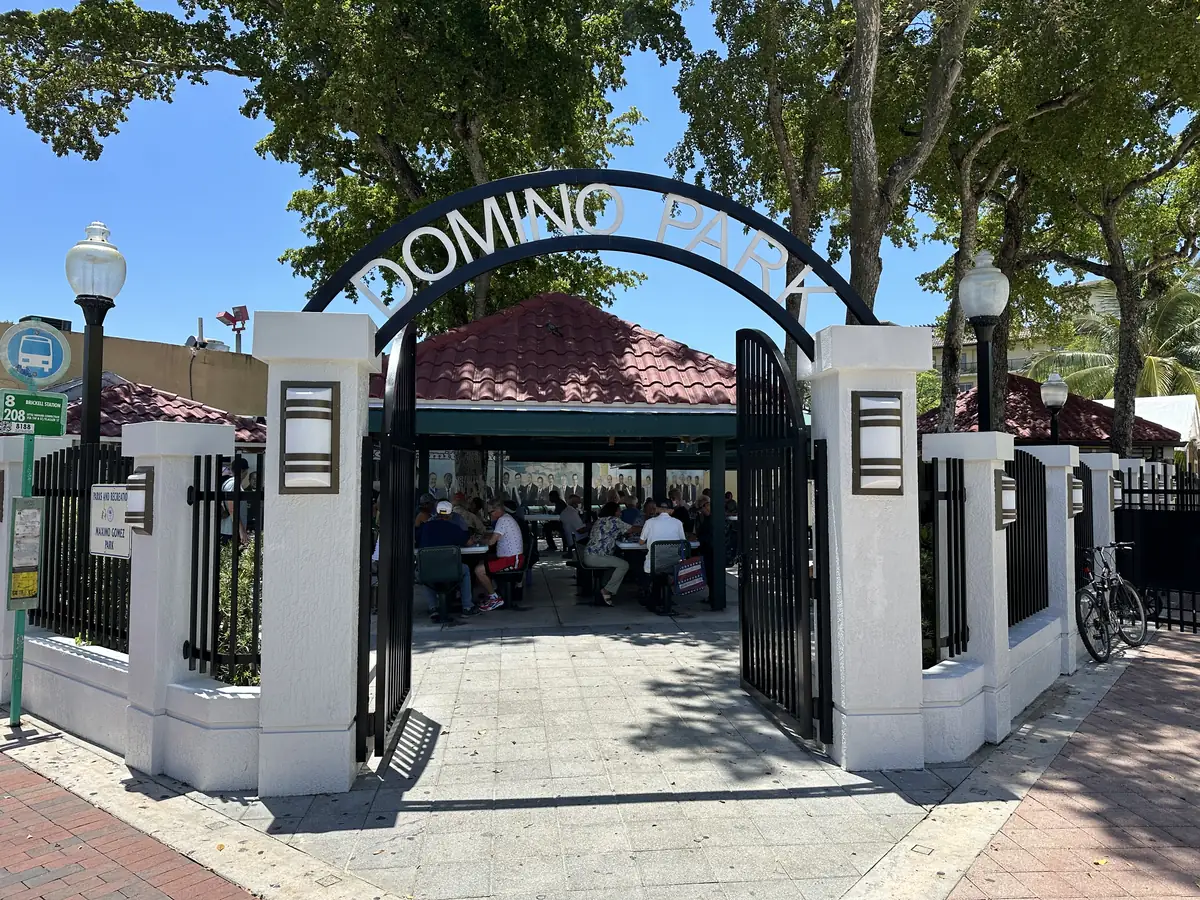A black iron arch reading Domino Park over an open gate between white pillars, with players at tables under a red tiled pavilion and shade trees behind
