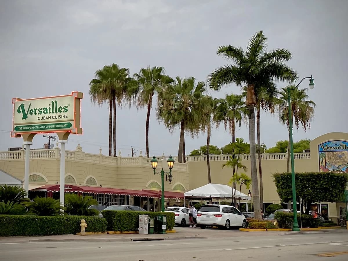 A green and cream Versailles sign on two posts in front of the long cream colored restaurant, with tall royal palms, street lamps and parked cars under a cloudy sky