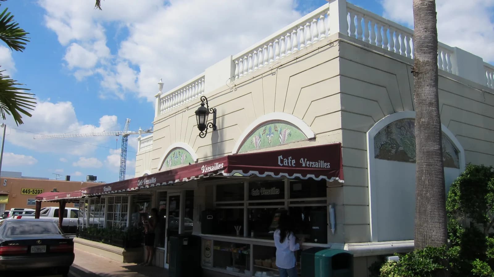 A cream colored restaurant with arched windows and a red awning over a walk up coffee window, with palm trees and blue sky