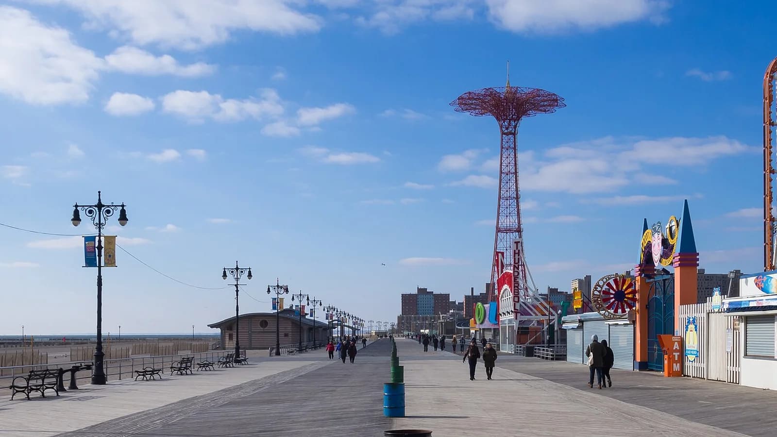 Riegelmann Boardwalk at Coney Island with lampposts, a few walkers and the red Parachute Jump tower under a blue sky
