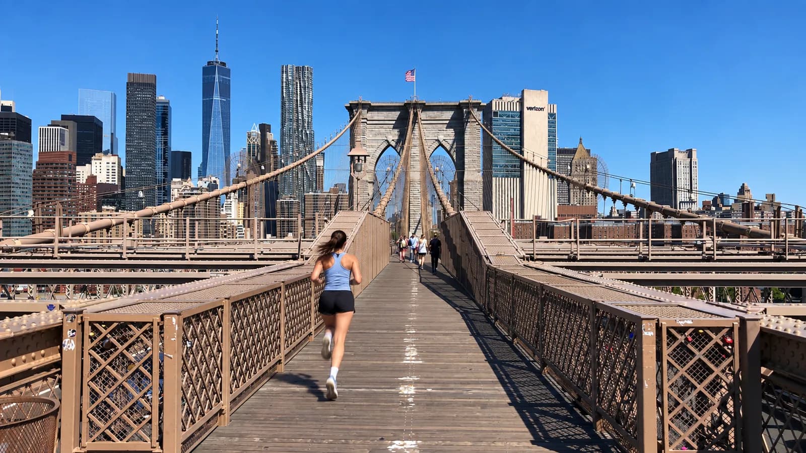 Woman in a light blue tank running the Brooklyn Bridge's wooden promenade toward the Manhattan tower and skyline