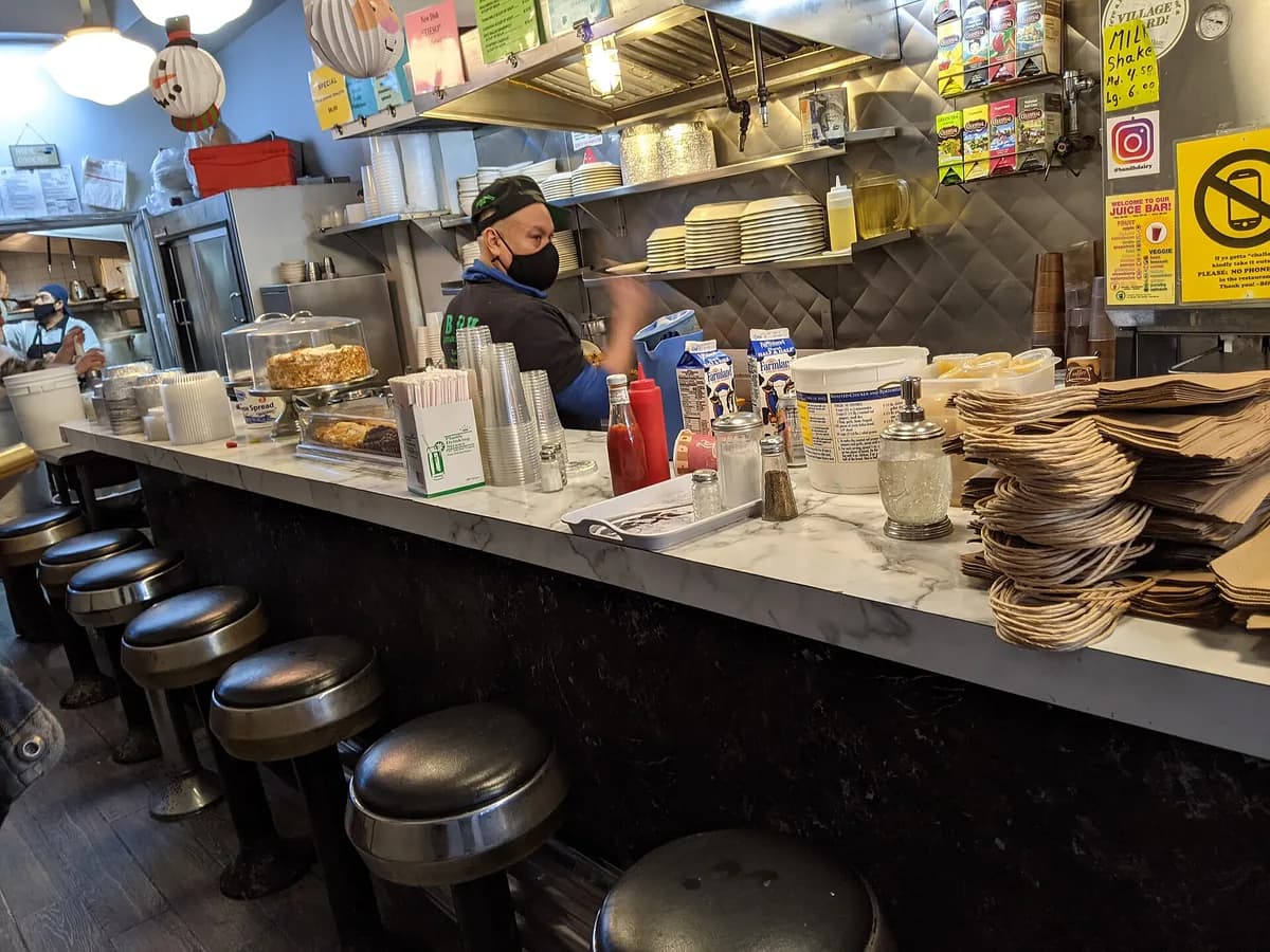 Long counter at B&H Dairy with a row of black stools, a cook at work behind it and stacked plates and supplies on the shelves