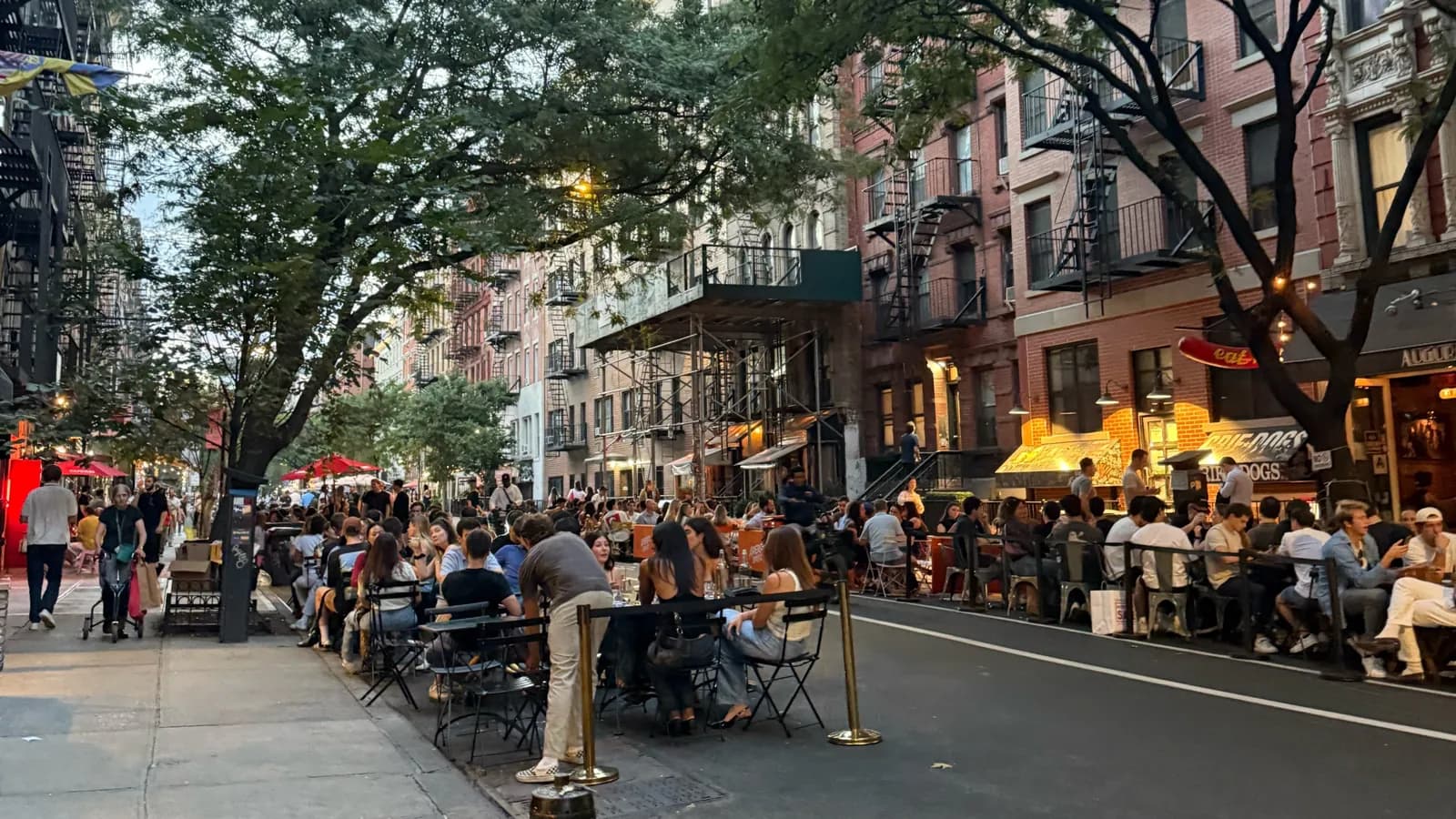 Outdoor dining tables filling the curb lane of tree-lined St. Marks Place in the East Village at dusk, with brick walk-ups and glowing shopfronts