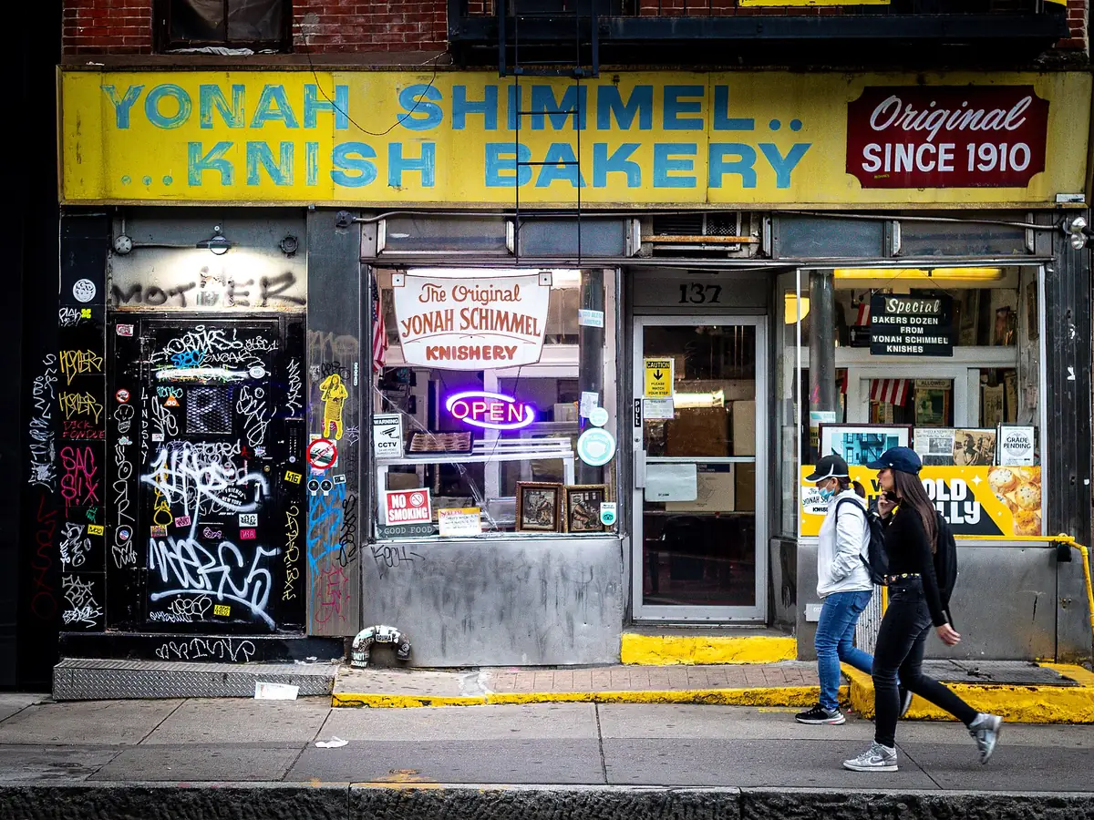 Yonah Schimmel Knish Bakery storefront on Houston Street with a yellow sign reading Since 1910, an old shop window and people walking past