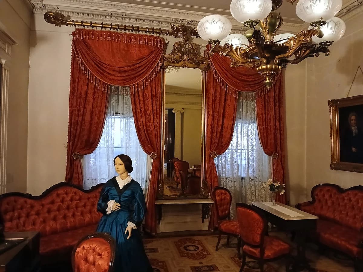 Front parlor of the Merchant's House Museum with red silk drapes, a gilt mirror, a brass gas chandelier, red tufted sofas and a costumed figure