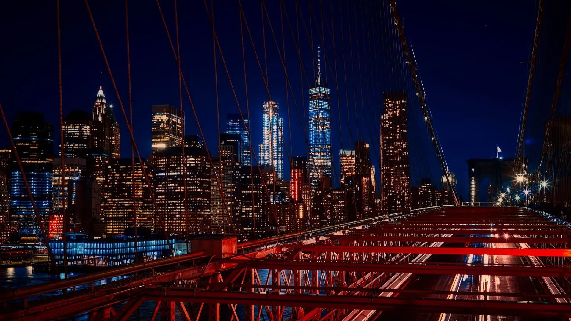 Lower Manhattan skyline and One World Trade Center lit up at night, seen through the cables of the Brooklyn Bridge