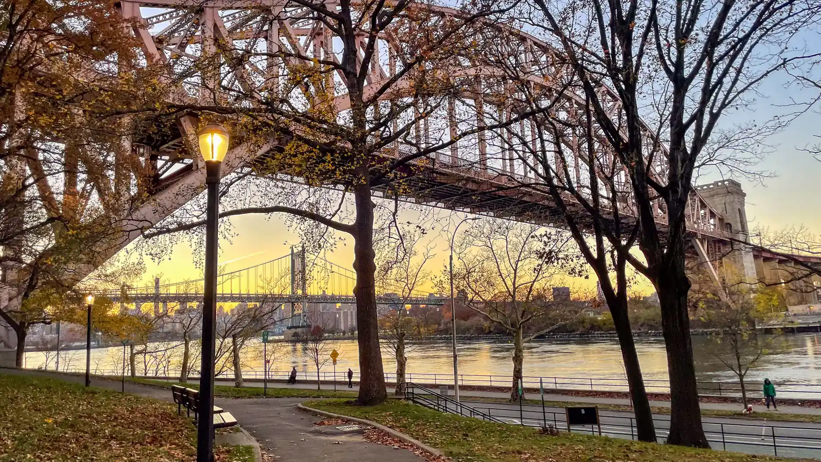 Steel arch of the Hell Gate Bridge over the East River seen from a tree lined path in Astoria Park at golden hour